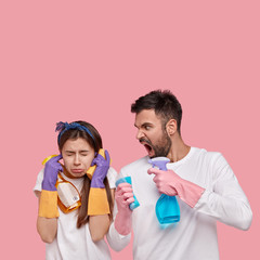 Fototapeta premium Outraged man with dark bristle screams at wife, reproaches of much dirt in kitchen, holds cleaning detergents. Sad woman ignores husbands scream, plugs ears, isolated on pink wall with empty space