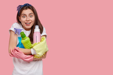 Studio shot of good looking pleased woman with dark straight hair, hugs cleaning detergents, wears headband and casual t shirt, isolated over pink background, blank space for your advertising