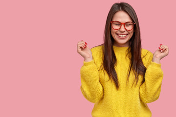 Happy joyful young woman Asian raises hands, smiles positively, wears spectacles, yellow loose...