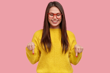 Charming happy young woman with gentle smile, points down with both index fingers, wears yellow clothes, enjoys something pleasant on floor, models against pink background. Promotion concept © WHstudio Leushin N