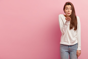 Mwah to you. Pleased attractive woman sends air kiss, has folded lips, wears casual pullover and jeans, expresses love to boyfriend, isolated over pink background with free space for your information
