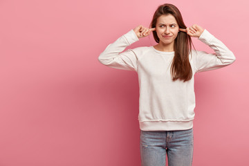 Indoor shot of dissatisfied woman plugs ears, hears awful noise, looks with unhappiness aside,...