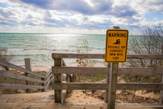 Rip Current Warning. Sign at beach entrance warning of dangerous rip currents as waves crash on to the coast of Lake Michigan.