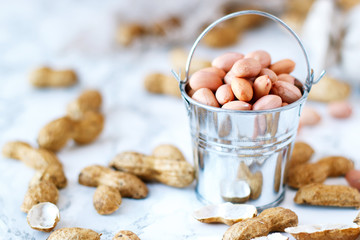 Raw peanuts in a bucket on a marble table. A peanut shell is lying next to a spoon.