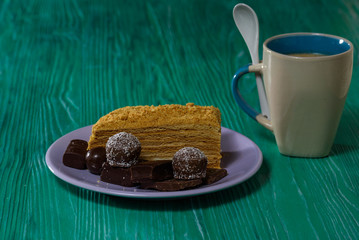 A piece of sponge cake and chocolates on a plate, next to it is a cup of coffee, photographed on a green wooden background close-up.