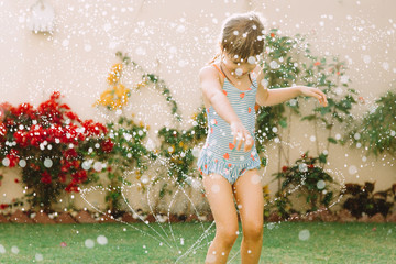 Funny laughing little girl  running though garden sprinkler playing with water splashes  in the backyard on a sunny hot summer vacation day . - obrazy, fototapety, plakaty