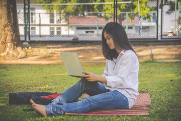 The asia woman is playing computer on a notebook.She sits and play laptop in park.