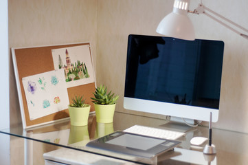 Glass desk with computer, graphic tablet, succulents in green pots and watercolor pictures on cork board. Modern desk workspace. Designer's workspace