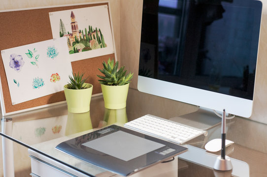Glass desk with computer, graphic tablet, succulents in green pots and watercolor pictures on cork board. Modern desk workspace. Designer's workspace
