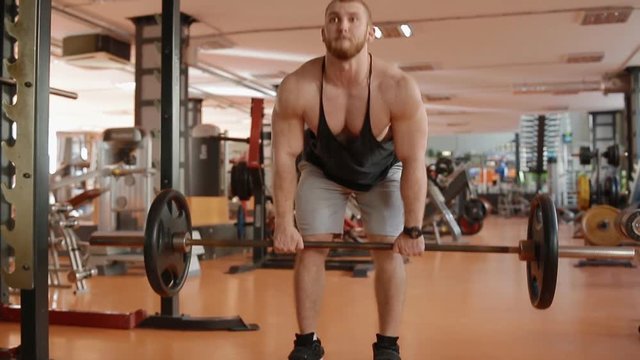 Young white bearded bodybuilder in black jersey making lifting of the barell to the belt. Emotion of pain on the face.