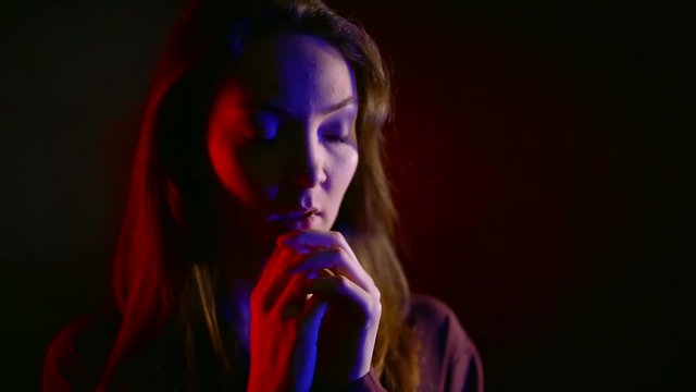 obedient young woman is holding hands folded near face, she is looking subordinately, standing in room