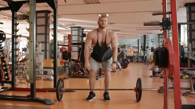 Young white bearded bodybuilder in black jersey making lifting of the barell to the belt. Emotion of pain on the face.
