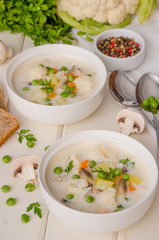 Cheese cream soup with cauliflower, mushrooms and green peas in a bowl on a white wooden background.