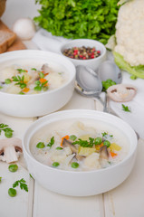 Cheese cream soup with cauliflower, mushrooms and green peas in a bowl on a white wooden background.