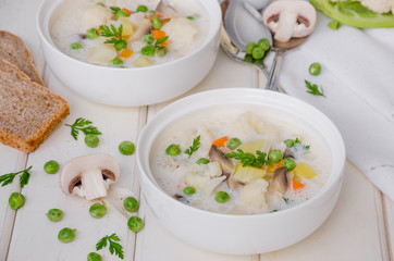 Cheese cream soup with cauliflower, mushrooms and green peas in a bowl on a white wooden background.