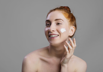 Close up beauty portrait of a young red hair and green eyes beautiful half naked woman applying face cream and looking at camera isolated over gray background