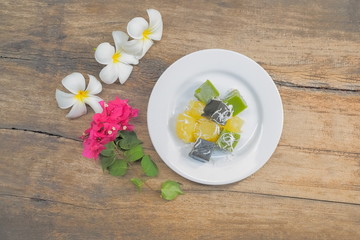 Delicious Thai Dessert Khanom Piek Poon (Burnt Coconut Jelly), Khanom Chun (Thai Jello) and Khanom Man (Coconut Cassava Cake) in white plate with flowers on wood texture background.