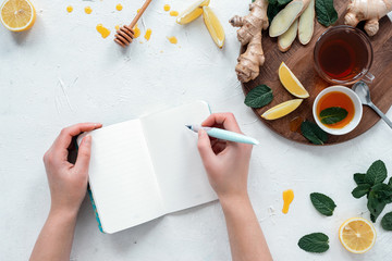 Young woman writes with ink pen in open notepad.Drink tea with ginger, lemon, mint, honey on white table.Top view.Healthcare concept.Tea against cold.