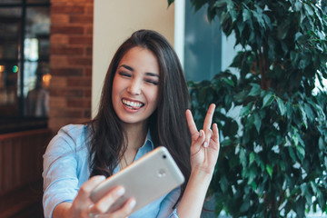 Beautiful charming brunette smiling asian girl taking selfie on frontal camera of smartphone at cafe
