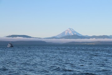 View of the Vilyuchinsky volcano (also called Vilyuchik) from tourist boat. The cloud lies on the coastal cliffs. Tourist boat is visible in the waters of Avacha Bay on the Kamchatka Peninsula, Russia