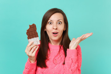 Surprised young woman in knitted pink sweater hold chocolate bar spreading hands keeping mouth wide...
