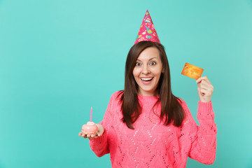 Surprised happy young woman in knitted pink sweater, birthday hat holding in hand cake with candle credit card isolated on blue turquoise wall background. People lifestyle concept. Mock up copy space.
