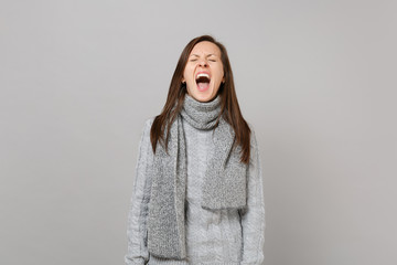 Weird young woman in gray sweater, scarf keeping eyes closed, mouth wide open screaming isolated on grey background. Healthy fashion lifestyle, people emotions cold season concept. Mock up copy space.
