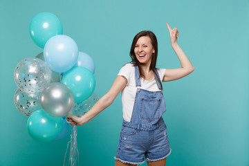 Laughing young woman in denim clothes pointing index finger up, celebrating, holding colorful air balloons isolated on blue turquoise wall background. Birthday holiday party, people emotions concept.