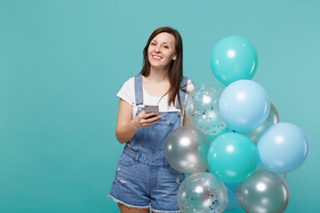 Fototapeta premium Beautiful smiling young woman in denim clothes holding mobile phone, celebrating with colorful air balloons isolated on blue turquoise wall background. Birthday holiday party, people emotions concept.