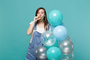 Fototapeta premium Amazed pretty young woman covering mouth with bitcoin, future currency celebrating holding colorful air balloons isolated on blue turquoise background. Birthday holiday party, people emotions concept.