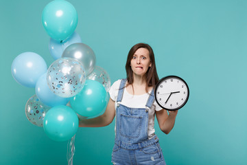 Concerned young woman in denim clothes biting lips hold round clock while celebrating with colorful...