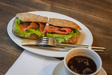 Fresh healthy sandwich with salmon and green lettuce in a white plate with cutlery, cup of coffee. Healthy diet food