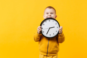 Little kid boy 3-4 years old wearing yellow clothes hold clock isolated on orange wall background,...