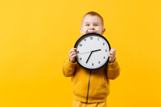 Little kid boy 3-4 years old wearing yellow clothes hold clock isolated on orange wall background, children studio portrait. People sincere emotions, childhood lifestyle concept. Mock up copy space.