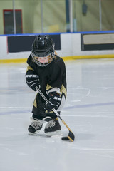 A little girl - hockey player on ice wearing in a full hockey equipment: helmet, black jersey, gloves with stick and puck.  