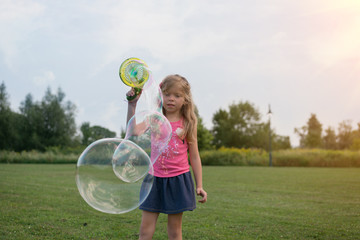 Adorable Caucasian blonde girl is making bubbles on the field during sunset in summer 