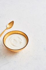 Obraz na płótnie Canvas Bowl of fresh Greek yoghurt and golden spoon on white abstract background. Healthy breakfast. Golden utensils. Top view. Copy space for text.
