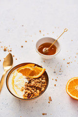 Bowl of homemade granola with yogurt, honey and fresh orange on white abstract background. Healthy breakfast. Golden utensils. Top view. Copy space for text.