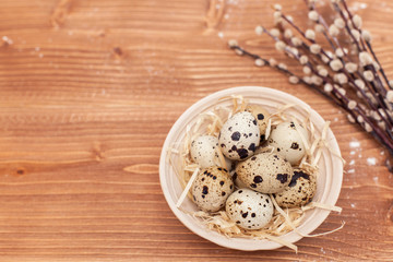 Happy Easter concept. Modern natural easter eggs on a wooden background with space for text. Quail eggs in a clay plate in the straw with willow branches. Flat lay