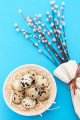 Happy Easter concept. Modern natural easter eggs on a blue background with space for text. Quail eggs in a clay plate in the straw with willow branches. Top view, vertical frame