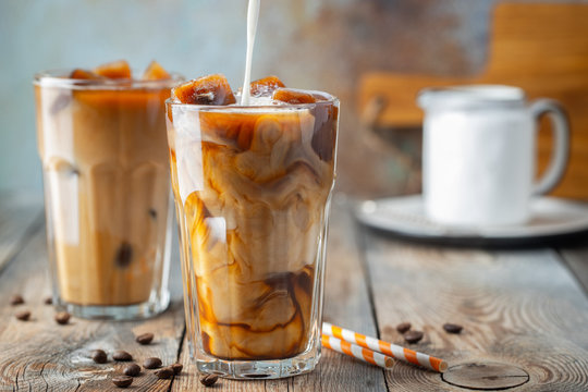 Ice coffee in a tall glass with cream poured over, coffee ice cubes and beans on a old rustic wooden table. Cold summer drink with tubes on a blue rusty background with copy space