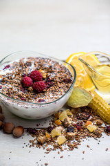transparent glass plate with muesli sprinkled with seeds and fresh raspberries stands on the table next to a mug of green tea, honeycomb and lemon slices