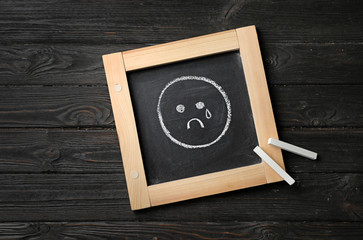 Small chalkboard with drawing of sad crying face on wooden background, top view. Depression symptoms