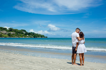 Fototapeta na wymiar Back view of young romantic couple in white clothes standing on tropical beach. Man and woman huging on left side of photo. Blue sky and see on background