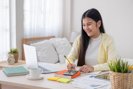 Asian female freelancer reading report on laptop and jot down note on table in bedroom at home.Work at home concept.work from home.relax lifestyle.