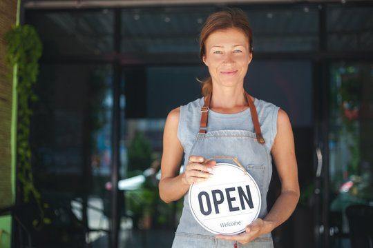 Woman waitress apron, cafe owner standing in the doorway, holding a sign with Open waiting for customers. Small business concept, cafes and restaurants