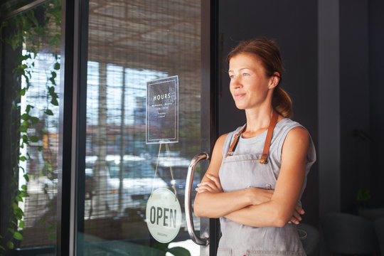 A female waitress in an apron, the owner of a cafe sits in the door of a cafe with a sign Open waiting for customers. Small business concept, cafes and restaurants