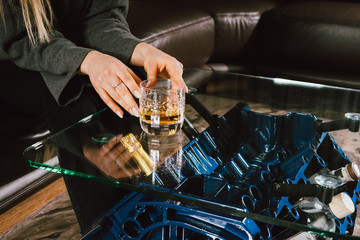 Young caucasian blonde girl sitting on sofa with glass of whiskey at luxury interior with custom v8  car engine table. Fashion picture and beautiful smile