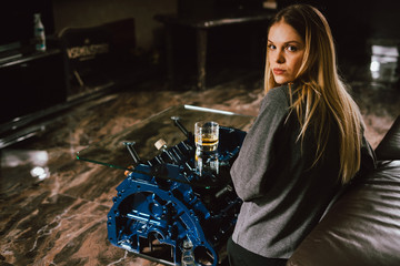 Naklejka na ściany i meble Young caucasian blonde girl sitting on sofa with glass of whiskey at luxury interior with custom v8 car engine table. Fashion picture and beautiful smile