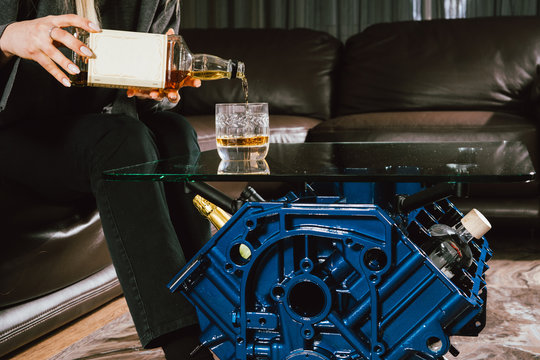 Young caucasian blonde girl sitting on sofa with glass of whiskey at luxury interior with custom v8  car engine table. Fashion picture and beautiful smile